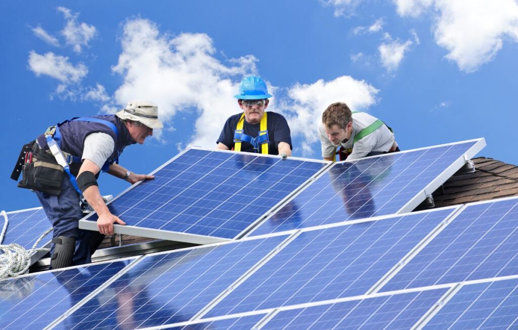 Workers install solar panels on a sunny rooftop. They are wearing safety harnesses and helmets while handling large blue panels under a bright sky. The image highlights renewable energy installation and sustainable practices.