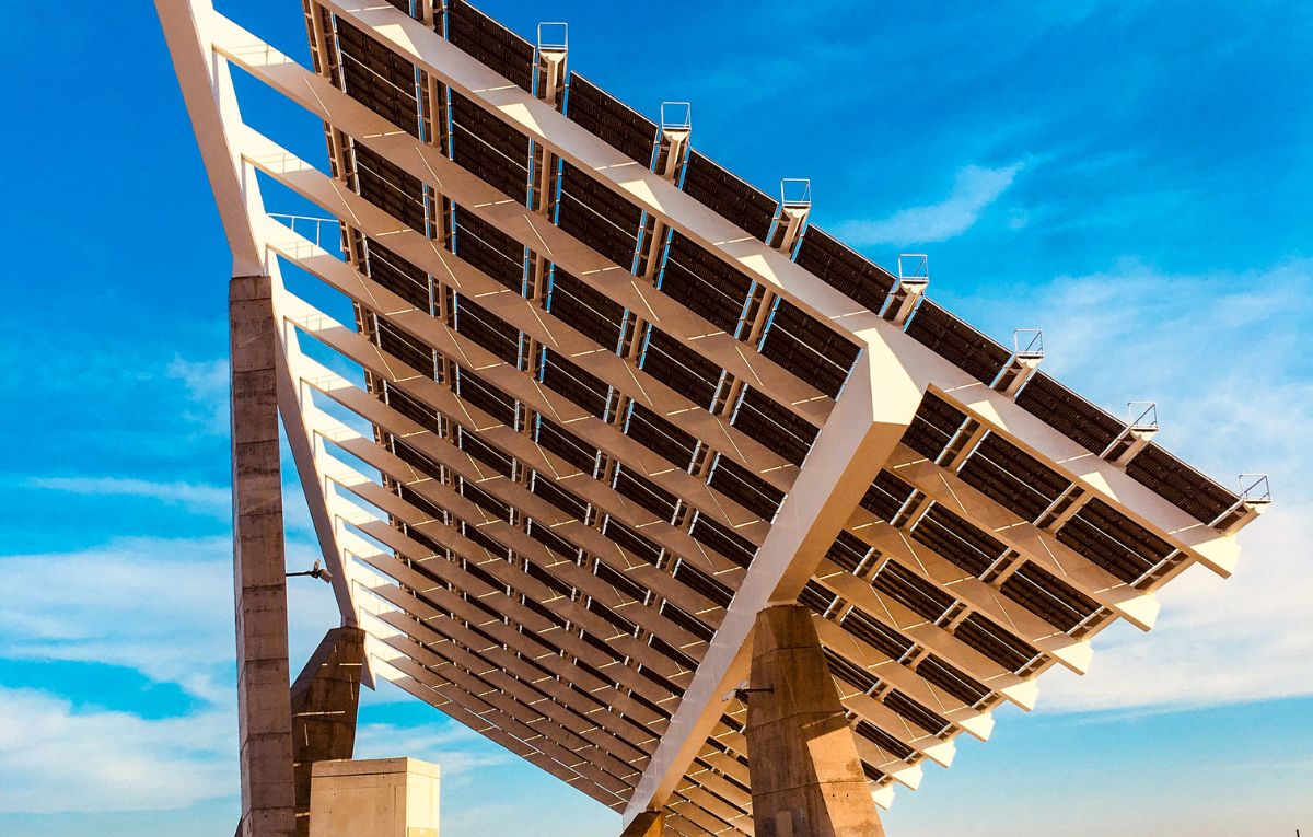 A large solar panel installation with multiple rows of panels mounted on a tilted frame supported by concrete pillars. The panels are arranged in a grid pattern and angled to maximize sunlight exposure. The background features a clear sky with a few clouds, indicating a sunny day ideal for solar energy generation.