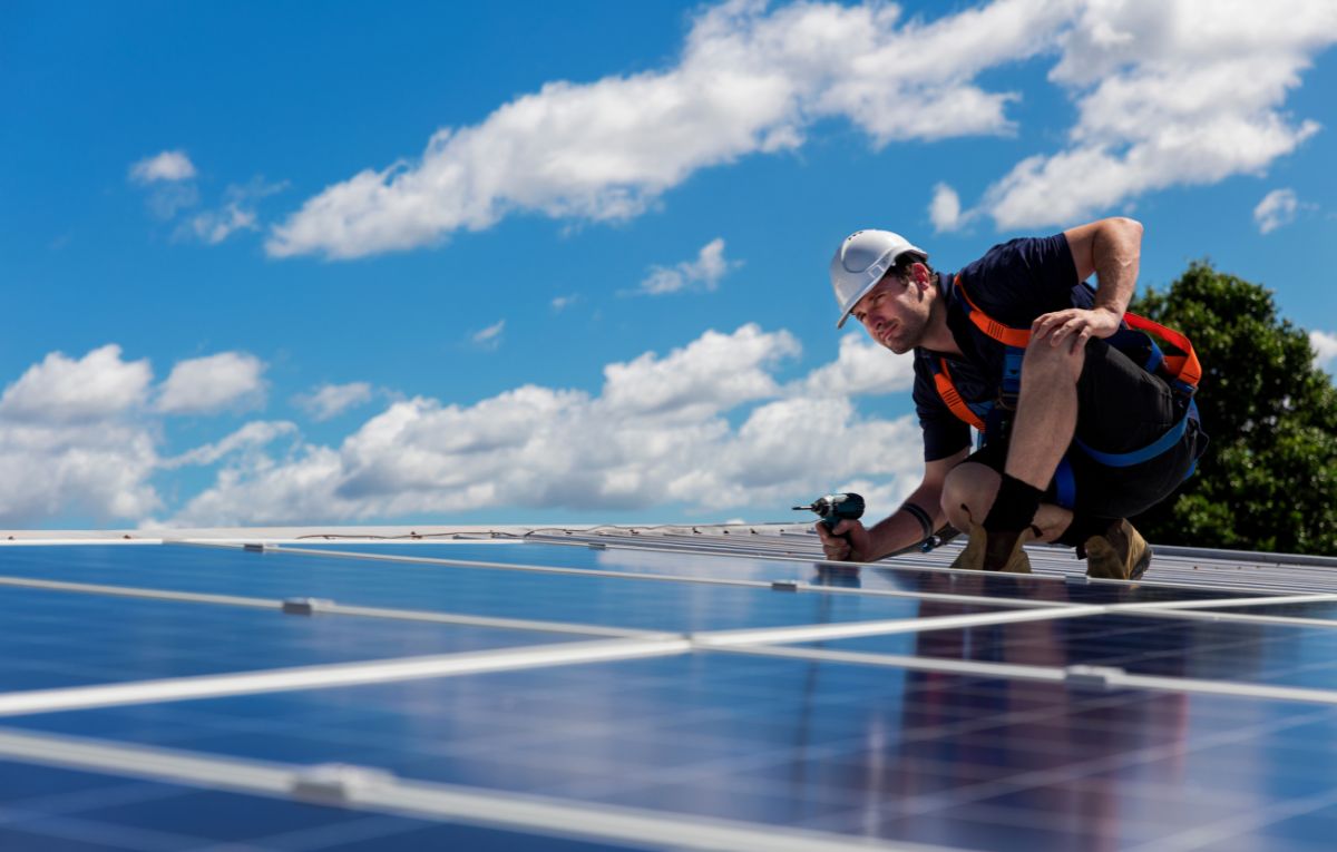 A solar panel installer awt the top of a roof with cottony clouds and blue sky at the background.