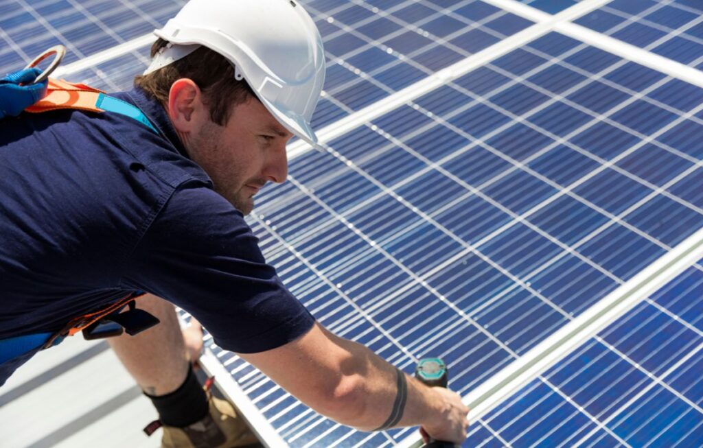 Technician wearing a white safety helmet and harness installing solar panels on a rooftop using a power drill, emphasizing renewable energy and professional solar system setup.